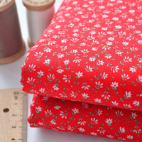 Two pieces of Longan Craft Floral Lawn Fabric By The Yard, featuring a small white floral pattern on red, are stacked beside a ruler and two spools of thread. This lightweight cotton lawn fabric is ideal for breathable dress or lining projects.