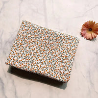A piece of Longan Craft Floral Printed Cotton Crepe Fabric By The Yard, featuring a delicate botanical pattern in small orange florals, is displayed folded on a white marble surface beside an orange-pink daisy flower.