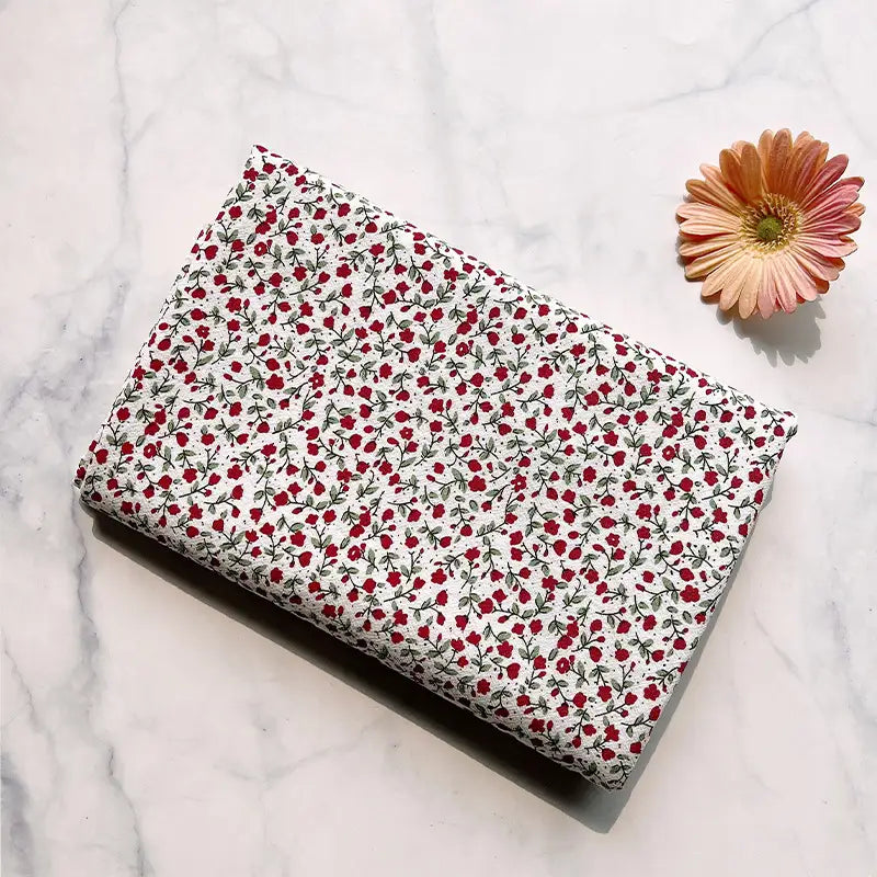A piece of Longan Craft Floral Printed Cotton Crepe Fabric, featuring a delicate washed crepe material with a botanical red and green pattern, is folded on a marble surface beside a single pink and yellow daisy.