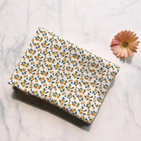 A piece of Longan Craft’s Floral Printed Cotton Crepe Fabric By The Yard, featuring a delicate botanical pattern in white with small yellow flowers, is folded and displayed on a marble surface beside a single pink and yellow daisy flower.