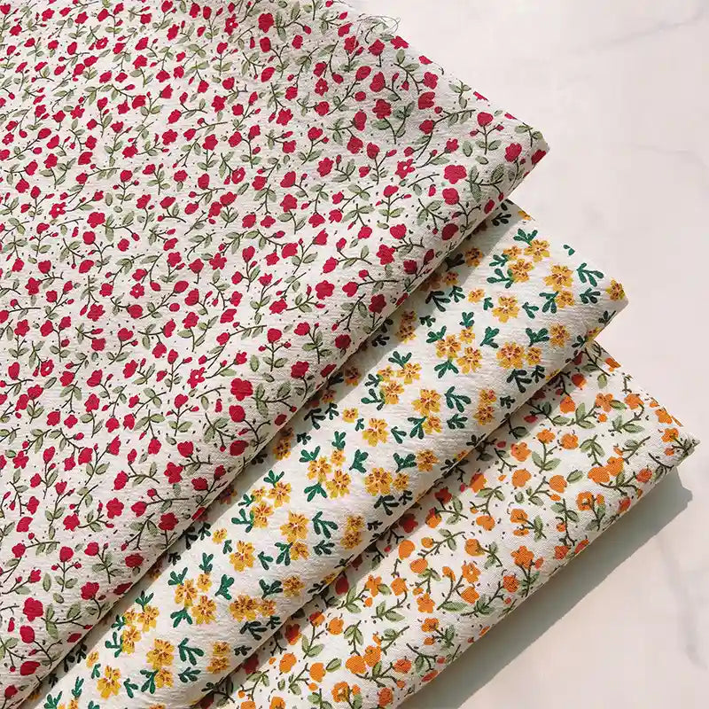 Four folded Longan Craft Floral Printed Cotton Crepe Fabrics are neatly arranged on a pale surface: the top fabric is a delicate cotton crepe printed with small red florals; beneath it lies a washed crepe material adorned with yellow flowers, followed by another featuring orange floral botanical patterns—all set on light backgrounds with green leaves, perfect for comfy dresses.