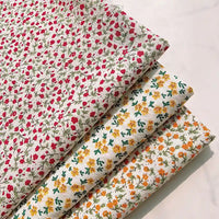 Four folded Longan Craft Floral Printed Cotton Crepe Fabrics are neatly arranged on a pale surface: the top fabric is a delicate cotton crepe printed with small red florals; beneath it lies a washed crepe material adorned with yellow flowers, followed by another featuring orange floral botanical patterns—all set on light backgrounds with green leaves, perfect for comfy dresses.