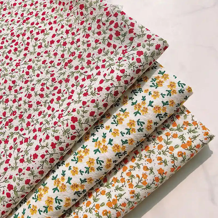 Four folded Longan Craft Floral Printed Cotton Crepe Fabrics are neatly arranged on a pale surface: the top fabric is a delicate cotton crepe printed with small red florals; beneath it lies a washed crepe material adorned with yellow flowers, followed by another featuring orange floral botanical patterns—all set on light backgrounds with green leaves, perfect for comfy dresses.