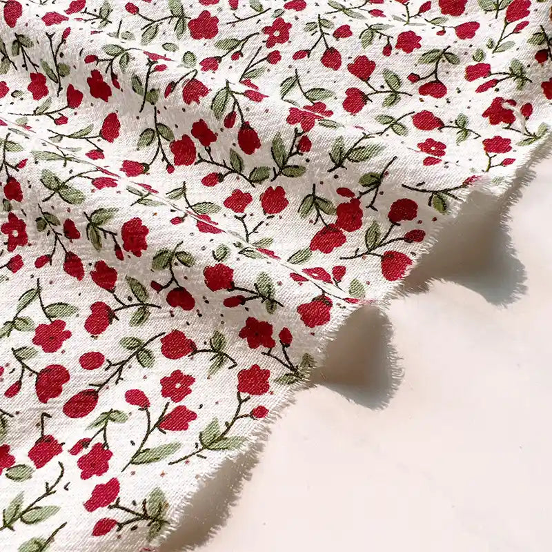 Longan Craft Floral Printed Cotton Crepe Fabric by the yard features a washed crepe material with a botanical pattern of small red flowers and green leaves, showcasing slightly frayed edges and gentle folds for added texture—perfect for comfy dresses.