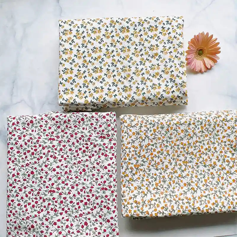 Three pieces of Longan Craft’s Floral Printed Cotton Crepe Fabric By The Yard, featuring botanical patterns in white, yellow, and red, are arranged on a white surface with a pink daisy flower placed nearby.