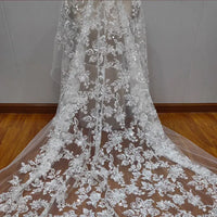 A detailed view of a white lace wedding dress train made with Longan Craft’s Floral Sequin Embroidered Tulle Fabric By The Yard, featuring intricate sequin and flower embroidery, spread over a wooden floor against a light wall.