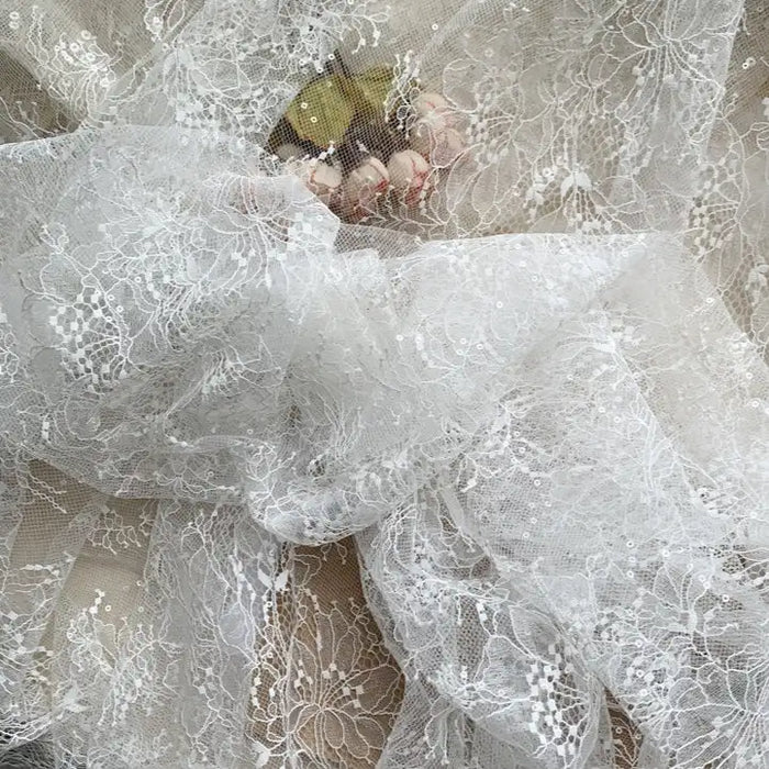 A hand holds Longan Craft's Floral Sequin Lace Fabric By The Yard, a colorful sparkling lace material for dresses, with delicate floral patterns and shimmering sequins partially covering the gold object.