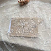 A beige envelope with a detailed overlay of Longan Craft's Floral Sequin Lace Fabric rests on a lace tablecloth, with pale pink flowers in the background. This colorful sparkling lace material is ideal for dresses and available by the yard.