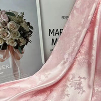A close-up of Longan Craft Floral Polyester Satin Fabric By The Yard in light pink, elegantly draped over a magazine page showing a bouquet of white and pale pink roses tied with a ribbon.