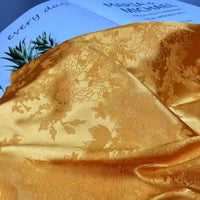 A piece of Floral Polyester Satin Fabric By The Yard from Longan Craft, featuring a colorful jacquard design, is draped on an open magazine with green leaves and partially visible text in the background.