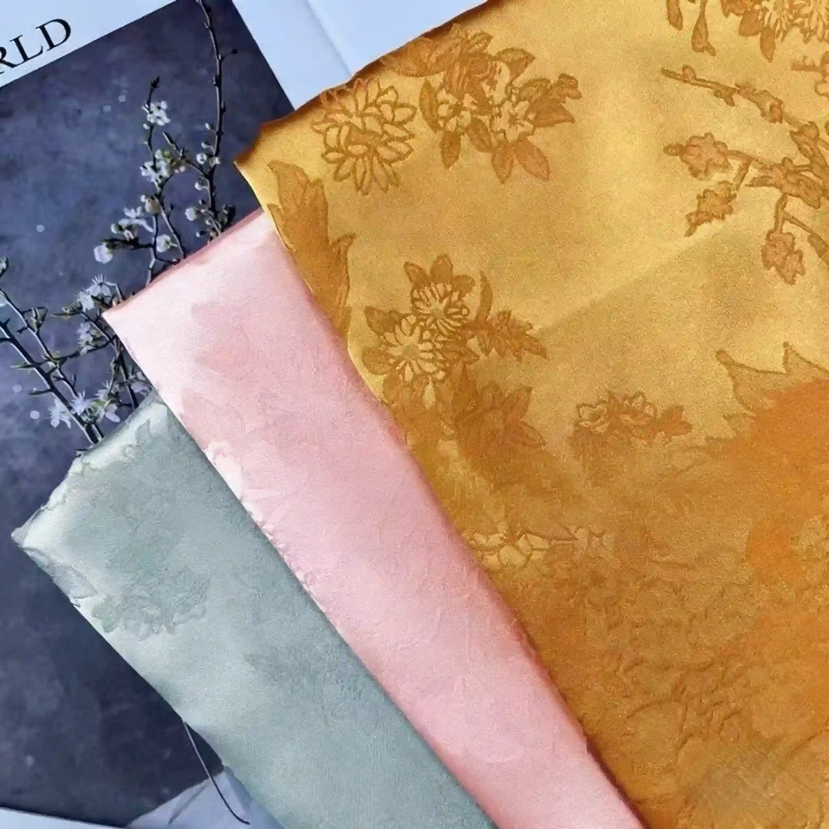 Three folded pieces of Longan Craft Floral Polyester Satin Fabric by the yard in pastel green, light pink, and golden yellow, each with subtle floral jacquard patterns, are displayed atop a magazine with a delicate branch and white blossoms nearby.