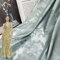 A woman in a gold, floral-patterned gown stands beside Longan Craft’s Floral Polyester Satin Fabric By The Yard — a colorful satin jacquard material for dresses. Behind it are a glass vase with flowers and an open book or magazine.