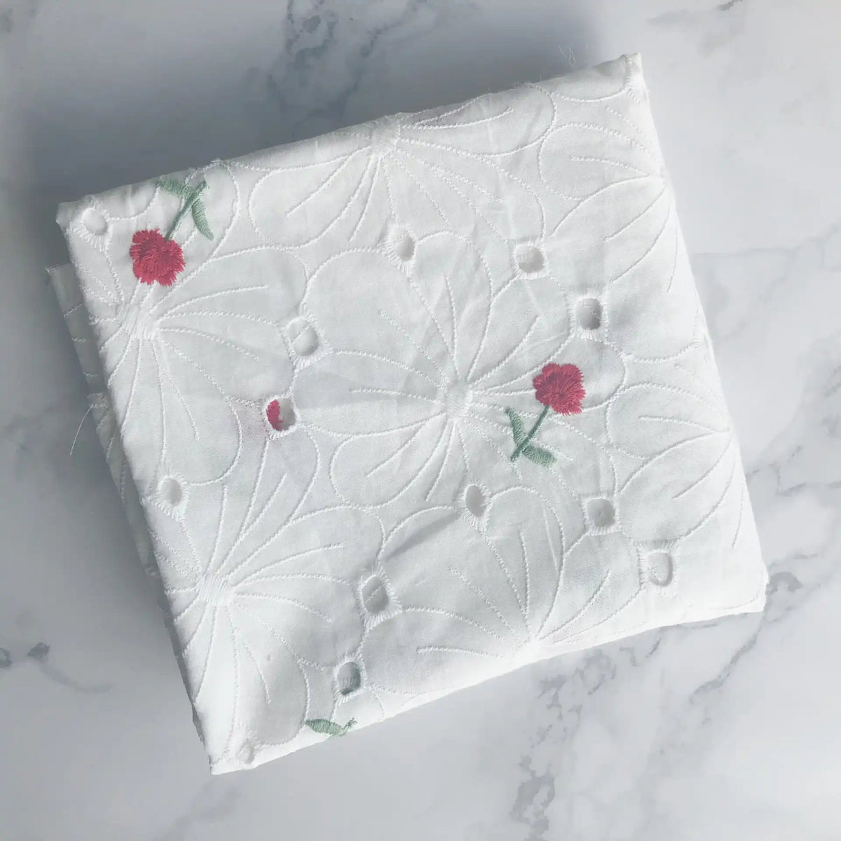 A folded piece of Longan Craft Flower Embroidered Cotton Eyelet Fabric in white, featuring delicate pink floral embroidery, is displayed on a light marble surface. Ideal for dresses, this soft cotton eyelet fabric is sold by the yard.