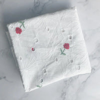 A folded piece of Longan Craft Flower Embroidered Cotton Eyelet Fabric in white, featuring delicate pink floral embroidery, is displayed on a light marble surface. Ideal for dresses, this soft cotton eyelet fabric is sold by the yard.