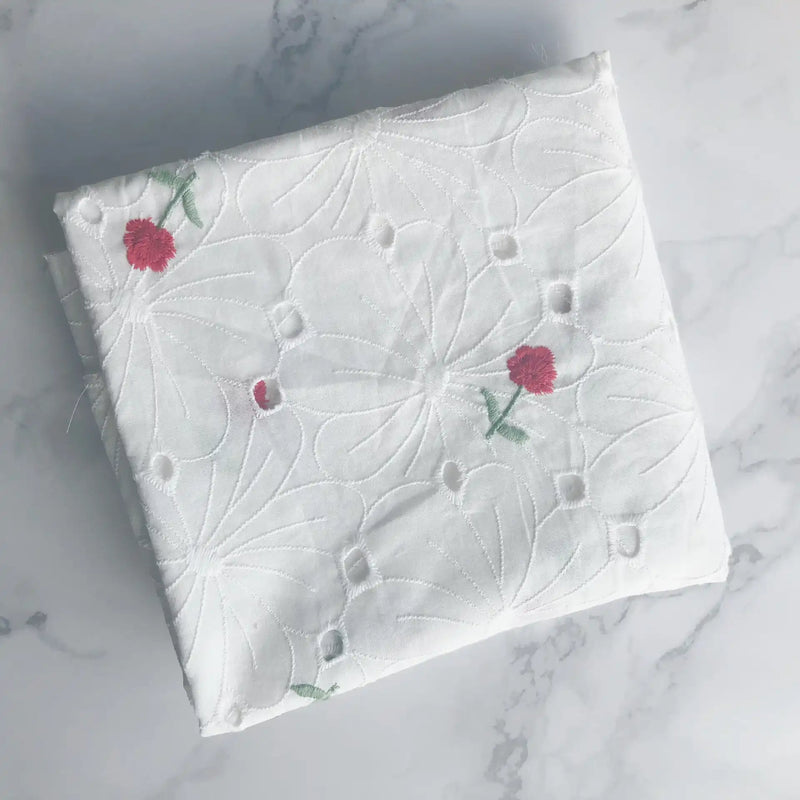 A folded piece of Longan Craft Flower Embroidered Cotton Eyelet Fabric in white, featuring delicate pink floral embroidery, is displayed on a light marble surface. Ideal for dresses, this soft cotton eyelet fabric is sold by the yard.