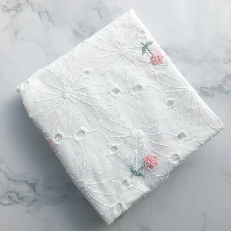 A neatly folded piece of Flower Embroidered Cotton Eyelet Fabric by Longan Craft, featuring delicate pink flowers and green leaves on white cotton, is displayed on a light marble surface. Ideal for dresses and sold by the yard.