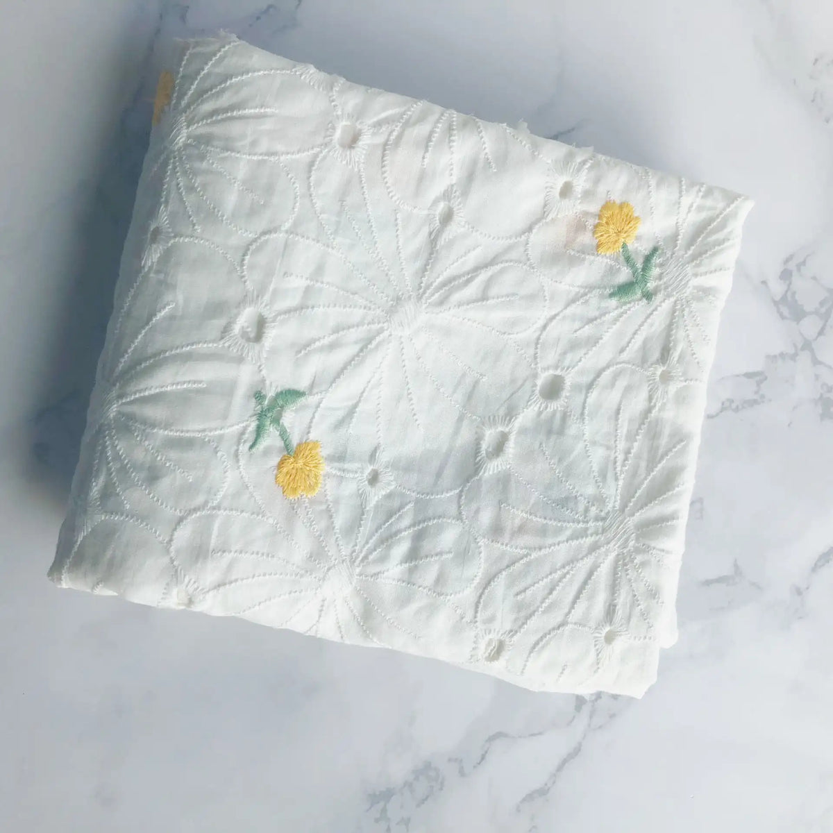 A folded piece of Longan Craft’s Flower Embroidered Cotton Eyelet Fabric by the yard, featuring white cotton eyelet material with floral embroidery, is displayed on a white marble surface.