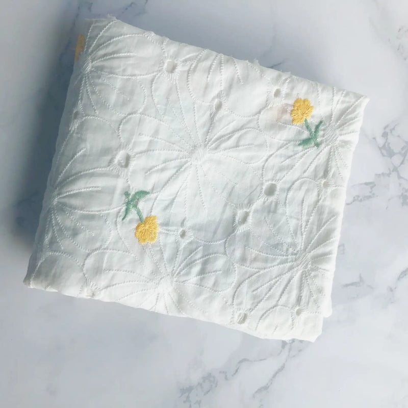 A folded piece of Longan Craft’s Flower Embroidered Cotton Eyelet Fabric by the yard, featuring white cotton eyelet material with floral embroidery, is displayed on a white marble surface.