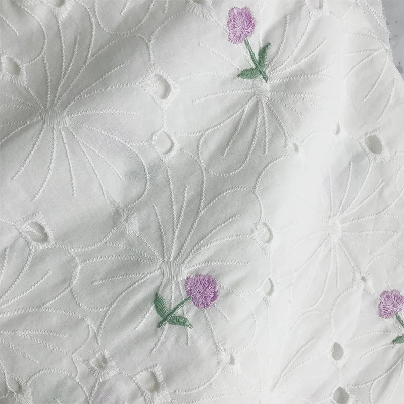 Longan Craft's Flower Embroidered Cotton Eyelet Fabric features white cotton material with large embroidered white flowers and delicate purple blooms with green stems, perfect for dresses and available by the yard.