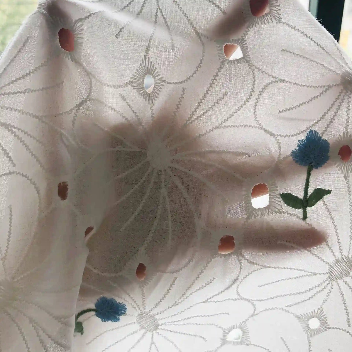 A hand is visible behind Longan Craft’s Flower Embroidered Cotton Eyelet Fabric By The Yard, a soft white cotton material with delicate blue floral embroidery and small cutouts that let light filter through—ideal for dresses.