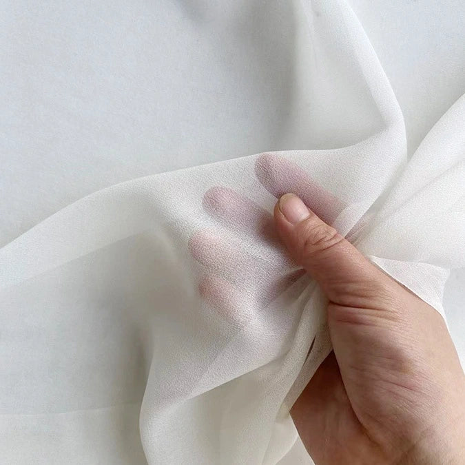 A hand pinches the sheer Georgette Silk Fabric By The Yard from Longan Craft, made of 8 Momme pure mulberry silk crepe, showing the soft, delicate texture and transparency against a plain background.