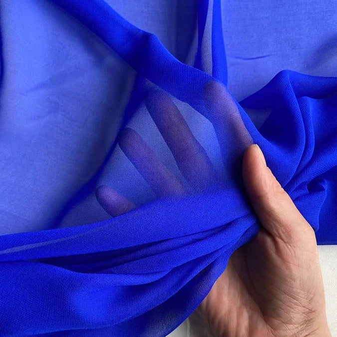 A hand holds up Longan Craft's vibrant blue Georgette Silk Fabric By The Yard—8 momme pure mulberry silk crepe that's sheer, smooth, and lightweight, with fingers visible through the translucent material.