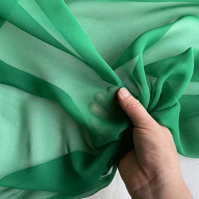 A hand gently holds a piece of Longan Craft’s Georgette Silk Fabric By The Yard, 8 Momme Pure Mulberry Silk Crepe, highlighting its soft texture and sheer green translucency as light reveals varied vibrant shades.