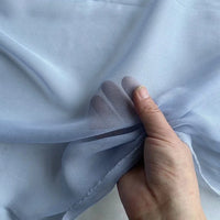 A hand holds up Longan Craft's Georgette Silk Fabric By The Yard—8 Momme Pure Mulberry Silk Crepe—showing fingers beneath its sheer, light blue, transparent texture.