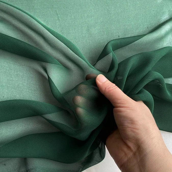 A hand holds a piece of Longan Craft Georgette Silk Fabric By The Yard (8 Momme Pure Mulberry Silk Crepe) against a green background, showing its lightweight, soft drape and slight transparency.