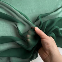 A hand holds a piece of Longan Craft Georgette Silk Fabric By The Yard (8 Momme Pure Mulberry Silk Crepe) against a green background, showing its lightweight, soft drape and slight transparency.