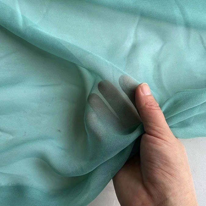 A hand holds Longan Craft's Georgette Silk Fabric By The Yard—8 Momme Pure Mulberry Silk Crepe—showing its sheer, light turquoise transparency with fingers visible through the soft drapery material.