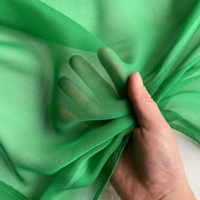 A hand bunches a piece of Georgette Silk Fabric By The Yard by Longan Craft, showing fingers through the sheer, green 8 momme pure mulberry silk crepe material.