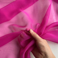 A hand gently gathers bright pink Longan Craft Georgette Silk Fabric By The Yard—8 Momme pure mulberry silk crepe—highlighting its soft, sheer, and delicate texture against a light background.