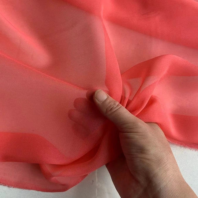 A hand holds a piece of Longan Craft Georgette Silk Fabric By The Yard—8 momme pure mulberry silk crepe—displaying its coral pink, sheer, delicate, and translucent texture against a light background.