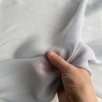 A hand gently lifts a piece of Longan Craft’s Georgette Silk Fabric By The Yard—8 Momme Pure Mulberry Silk Crepe—showcasing its soft, sheer, and delicate texture against a plain background.