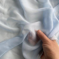 A hand gently lifts Longan Craft's Georgette Silk Fabric By The Yard—8 Momme pure Mulberry silk crepe—revealing its light blue, soft, and delicate texture.