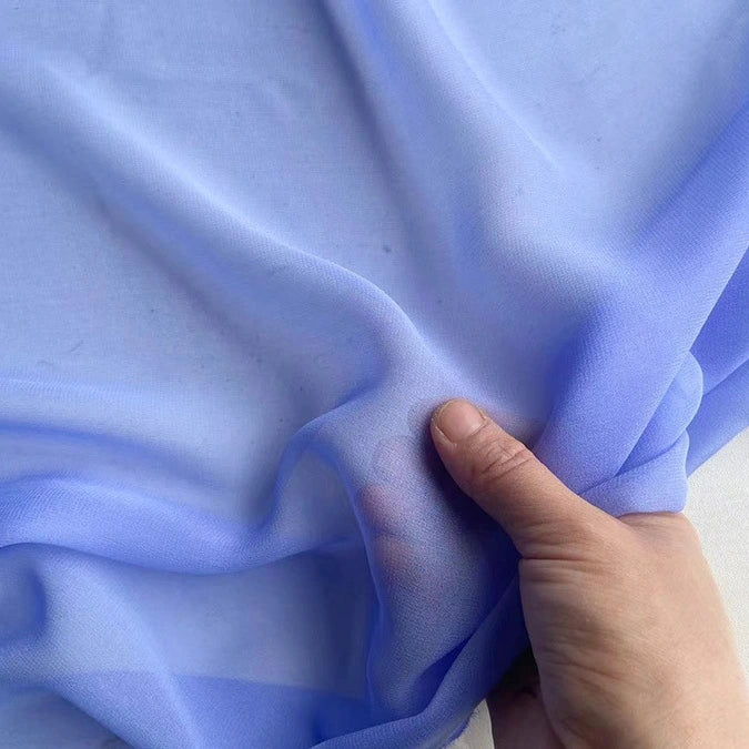 A hand gently gathers Longan Craft Georgette Silk Fabric By The Yard—8 Momme pure mulberry silk crepe—in sheer, light blue, against a neutral background.