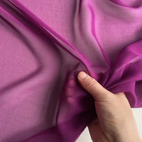 A hand gently gathers sheer, bright purple Longan Craft Georgette Silk Fabric by the Yard—8 momme pure mulberry silk crepe—showing its lightweight, translucent texture against a light background.