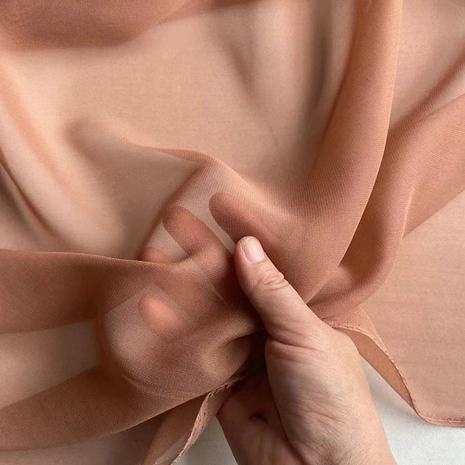 A hand gently holds a piece of Longan Craft Georgette Silk Fabric By The Yard, 8 Momme Pure Mulberry Silk Crepe, highlighting its soft, sheer texture and delicate translucence.