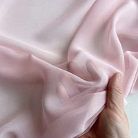 A hand gathers sheer, light pink Georgette Silk Fabric from Longan Craft, highlighting the soft, delicate texture and semi-transparency of this 8 Momme Pure Mulberry Silk Crepe Fabric by the yard—ideal for elegant drapery projects.