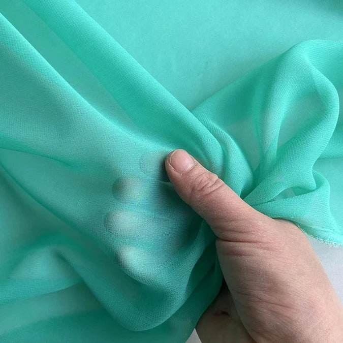 A hand lifts a piece of Longan Craft Georgette Silk Fabric By The Yard—8 Momme Pure Mulberry Silk Crepe—highlighting its delicate, sheer texture and soft drape against a matching background.