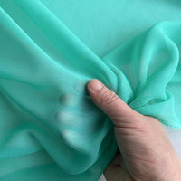 A hand lifts a piece of Longan Craft Georgette Silk Fabric By The Yard—8 Momme Pure Mulberry Silk Crepe—highlighting its delicate, sheer texture and soft drape against a matching background.