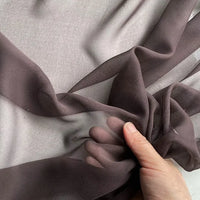 A hand gathers and wrinkles Longan Craft's Georgette Silk Fabric By The Yard—8 Momme pure mulberry silk crepe—creating soft folds and shadows on the sheer, dark brown drapery fabric against a light background.