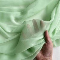 A hand lifts the sheer, light green Georgette Silk Fabric By The Yard from Longan Craft, made from 8 Momme Pure Mulberry Silk Crepe. The translucent drapery reveals finger outlines against a soft, neutral background.