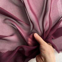 A hand gathers Longan Craft’s Georgette Silk Fabric By The Yard—8 Momme Pure Mulberry Silk Crepe—showing its wine hue, delicate texture, and semi-transparency as light highlights the soft, flowing folds of this drapery fabric.