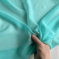 A hand gently holds and gathers a light turquoise Georgette Silk Fabric by Longan Craft, showcasing the 8 Momme pure mulberry silk crepe's delicate, translucent texture against a light background.
