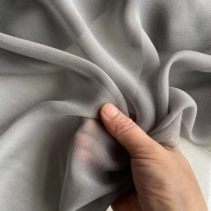A hand gently grasps Longan Craft’s Georgette Silk Fabric By The Yard—soft, flowing 8 Momme pure mulberry silk crepe—creating delicate folds and texture in the semi-transparent gray drapery material.