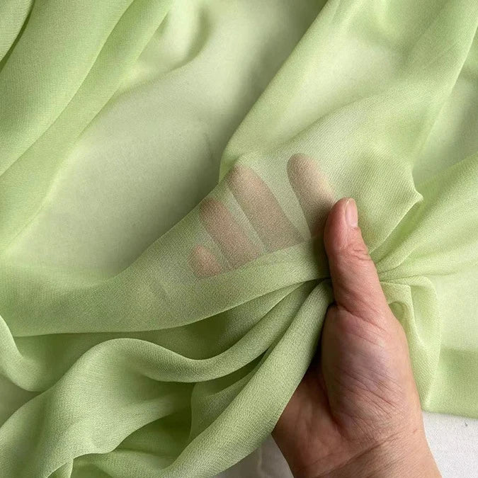 A hand holds a piece of Longan Craft Georgette Silk Fabric By The Yard—8 Momme Pure Mulberry Silk Crepe—its light green, sheer, soft silk texture revealing fingers through the translucent drapery material.