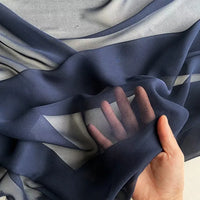 A hand lifts a piece of Longan Craft Georgette Silk Fabric By The Yard, 8 Momme Pure Mulberry Silk Crepe, revealing its soft, sheer texture and semi-transparency as light filters through both the fabric and fingers.