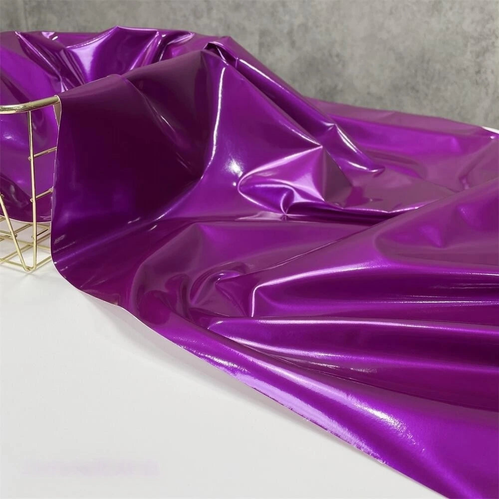 A yard of Longan Craft's Glossy Stretch Vinyl Fabric in purple is draped over a white surface, with a gold wire basket on the left and a gray textured background behind. This shiny PU vinyl is perfect for making jackets.